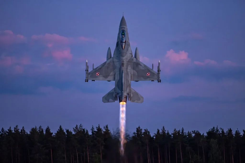 F-16 Jastrząb wznoszący się pionowo z jasnym płomieniem dopalacza nad ciemnym lasem w fioletowym zmierzchu.